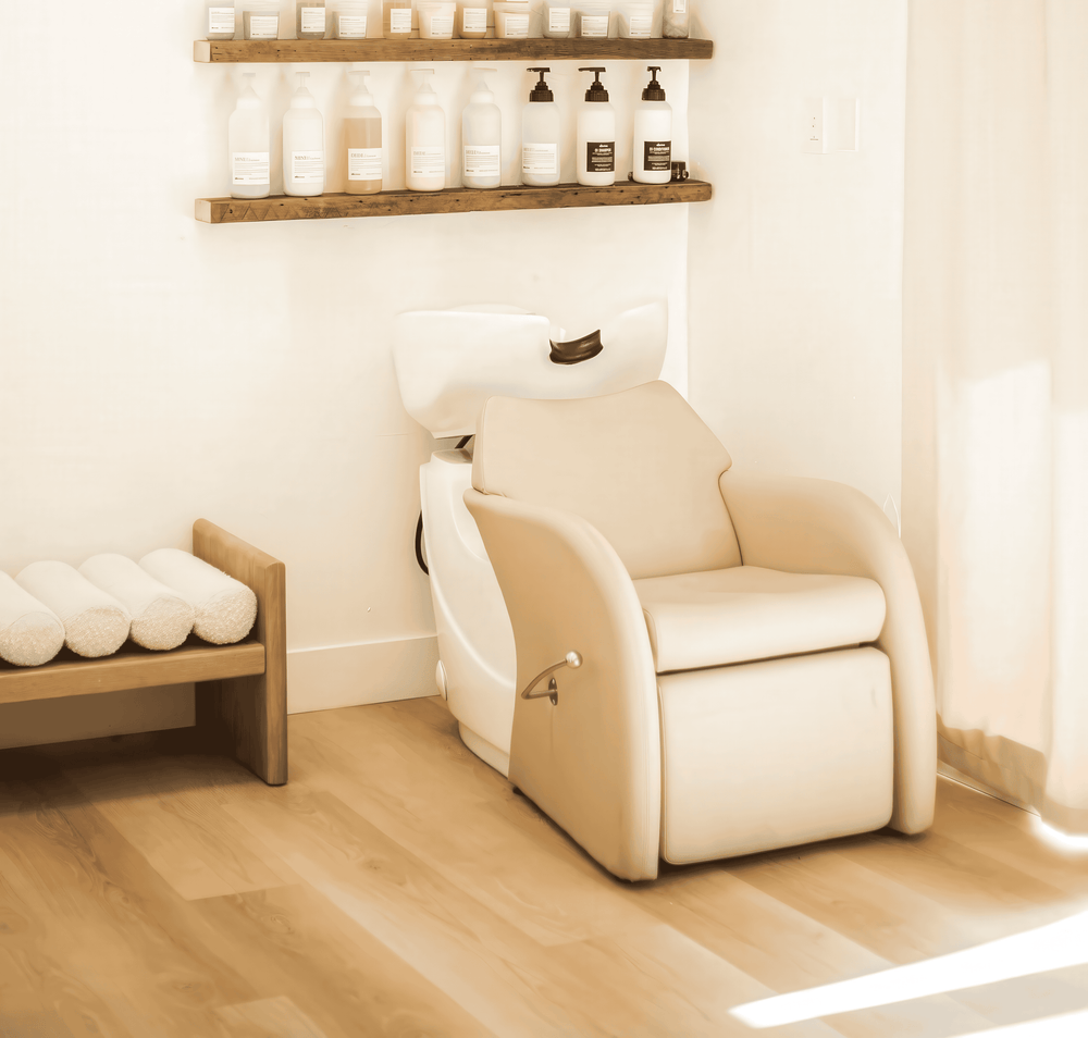 A cream-colored reclining spa shampoo chair, placed in a bright, minimalist spa room with light wood floors, a wooden shelf with bottles, and a towel bench.