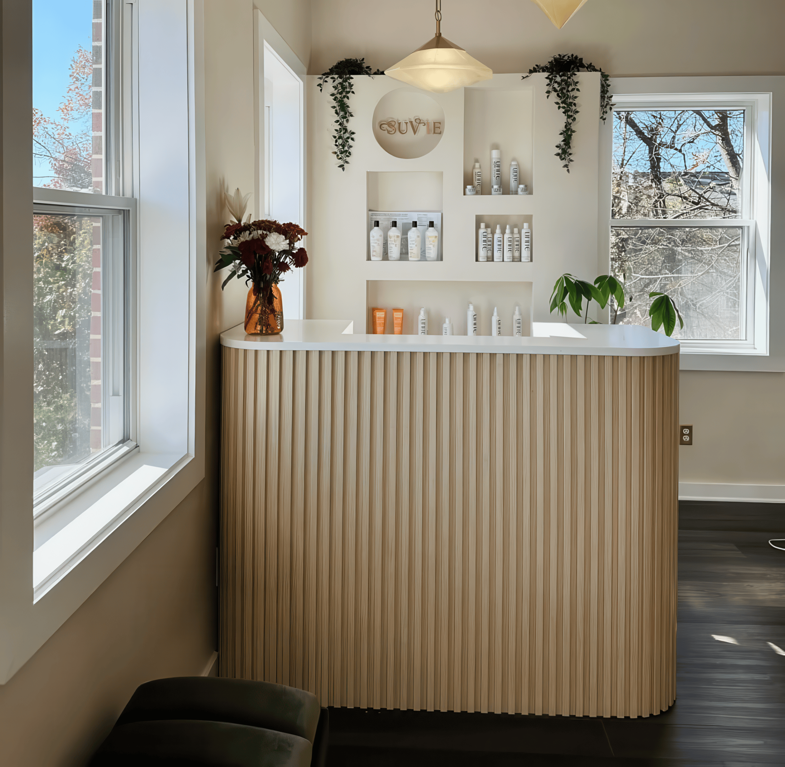 A modern interior design scene featuring a fluted light wood reception desk or vanity unit with a white countertop, set against a beige wall with built-in shelves and plants.