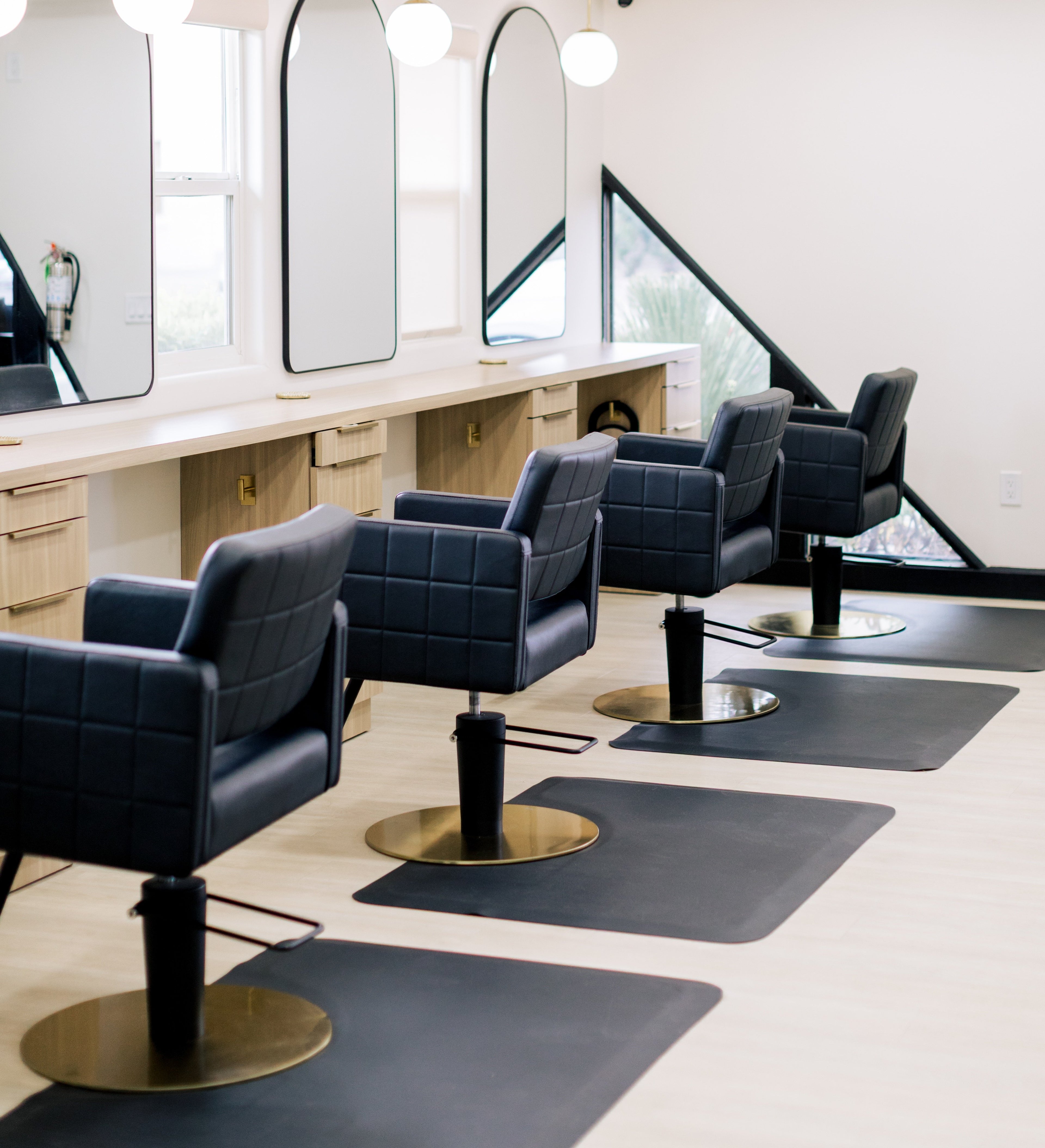 A row of modern black tufted salon chairs with gold bases arranged in a bright, contemporary hair salon with light wood counters and mirrors.
